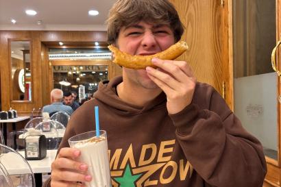 My friend Caden sits inside the churro shop at night, smiling. His face is scrunched up and he holds a curved churro in front of his mouth to mimic a smile, clutching a cinnamon-dusted leche rizada in the other hand.