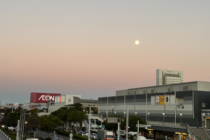 Picture of a sunset by a train station and road, the moon is full and the sky is a gradient of pale orange to blue