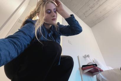 The author of the blog crouching on the floor and posing in a long sleeve denim shirt, black slacks, and white sunglasses. In the background, the Collegio dorm room is partially on display. You can see part of the grey ceiling and all white walls. 