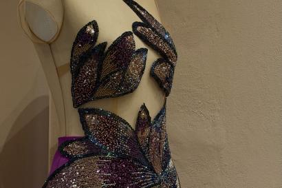 A photo of a gown with a sheer nude bodice and a purple skirt joined together with beaded appliqués that cover the bust and the waist. The beads are in gradients of gold, purple, and pink.   