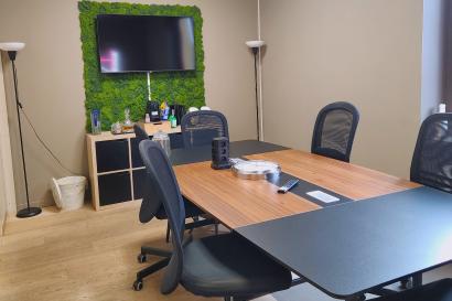 A small meeting room with a wooden conference table, office chairs, a wall-mounted TV, and a green moss accent wall
