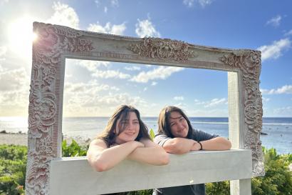 Two students smiling and posing in a large frame outside at the Cook Islands while the sun is shining behind them