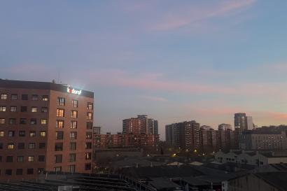A view of the sunset through a bedroom window in Aparto Cristobal. A tall red student living building is seen along with other buildings in the area.