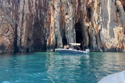 small boat floats in clear blue water and wall of reddish rocks behind it 