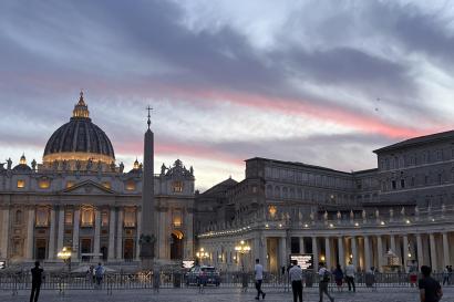 A view of St. Peter’s Basilica and St. Peter’s Square at sunset, with the basilica being illuminated by warm lights. 