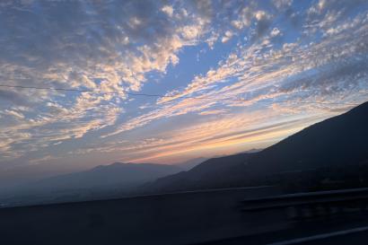 Soft yellows, warm oranges, and pale blues illuminate the cloud-filled sky behind misty silhouettes of mountains 