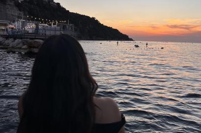 A person sits by the water in Sorrento, watching the sunset over the sea as the sky is pink and orange. 
