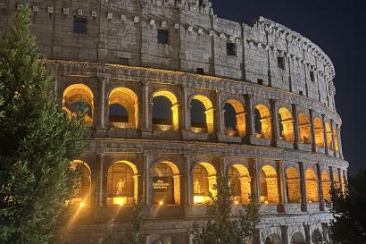  Illuminated view of the Colosseum in Rome at night, with its arches glowing yellow against the dark sky and trees in the foreground.