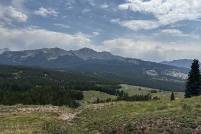 A mountain landscape, with multiple mountains and clouds in the background and greenery in the foreground.
