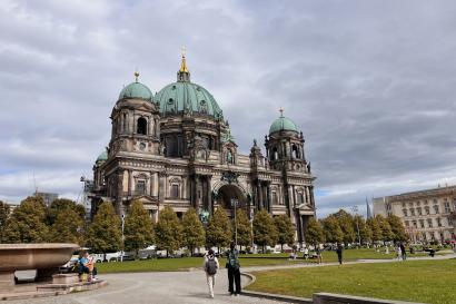 Two tourists stop to take photos in front of the Berlin Cathedral, a Baroque-style palace with a green center dome and four corner towers.