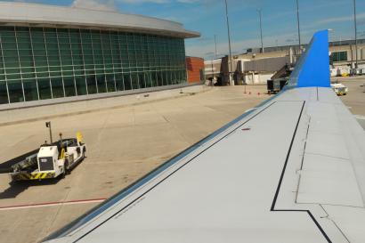 Airplane wing with a bright blue wingtip on an airport tarmac. The photo is taken from a window seat near the wing, showing the long white wing stretching toward the distance, with flap panels visible along its trailing edge. Below the wing is a concrete apron with a red guideline and some darker stained areas. Near the center-bottom of the image there is a small white and yellow ground vehicle positioned close to the wing, with a tow bar extended toward the aircraft. To the left side of the scene is an air