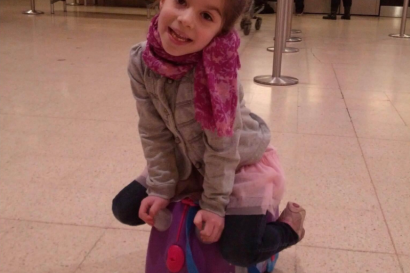 Young girl smiling while sitting on a small purple ride-on suitcase in an airport terminal.