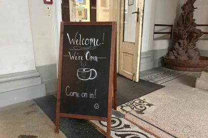 A sign on a clapboard reads "Welcome! We're open. Come on in" and has a cartoon coffee cup drawing. An open door is behind it.