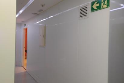 White hallway in a modern building. The corridor has smooth white walls and a white ceiling with recessed lighting running along it. On the right upper part of the image, there is a green emergency exit sign with a running figure and a large left-pointing arrow, indicating the exit is toward the left. Just to the left of that sign is a rectangular wall vent. Farther down the hallway on the left side, there is an open doorway; inside that room the walls or lighting appear orange, and a darker object is partl