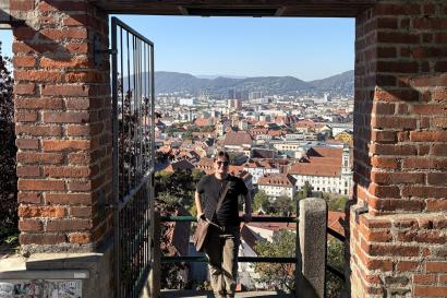 The author leans against a railing above a view of Graz, Austria, with many old buildings below. The photo is framed on both sides with brick pillars.