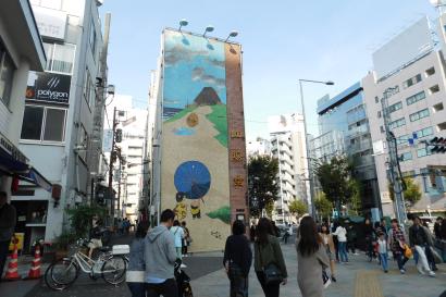Street with a large mural and people walking, tall buildings on either side. This alt text was added with Al; accuracy may vary.
