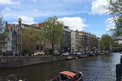 Canal with boats, lined with historic buildings and trees under a blue sky. This alt text was added with Al; accuracy may vary.