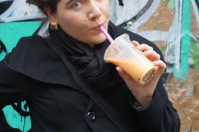 A person in a black coat drinks a beverage with graffiti in the background. This alt text was added with Al; accuracy may vary.