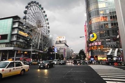 Bustling city street with Ferris wheel and skyscrapers under a cloudy sky. This alt text was added with Al; accuracy may vary.