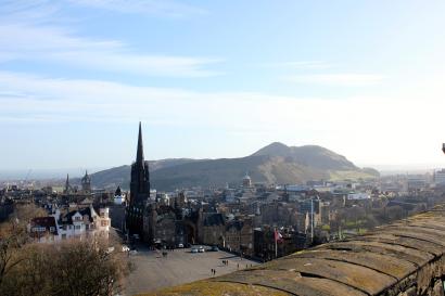 Cityscape view from a castle with a prominent spire and a hilly backdrop under a clear sky. This alt text was added with Al; accuracy may vary.