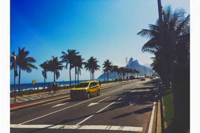 Avenida by the ocean lined with palm trees, yellow car driving, blue sky. This alt text was added with Al; accuracy may vary.