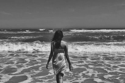 A woman wades in the ocean, wearing a dress, seen in black and white. This alt text was added with Al; accuracy may vary.