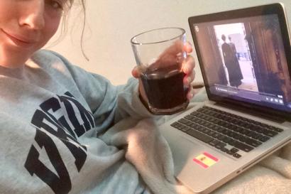 Person smiling, holding a drink, watching a show on a laptop. Cozy setting. This alt text was added with Al; accuracy may vary.