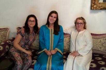 Three women in festive attire sitting on a patterned couch, smiling indoors. This alt text was added with Al; accuracy may vary.