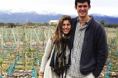 A couple stands in a vineyard with mountains in the background. This alt text was added with Al; accuracy may vary.