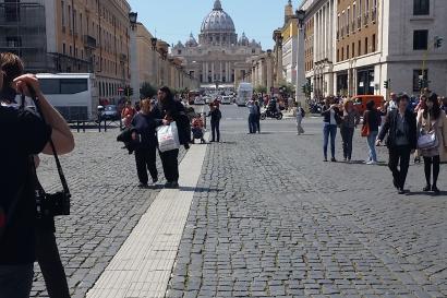 Wide street view towards St. Peter's Basilica, Rome, with people walking. This alt text was added with Al; accuracy may vary.
