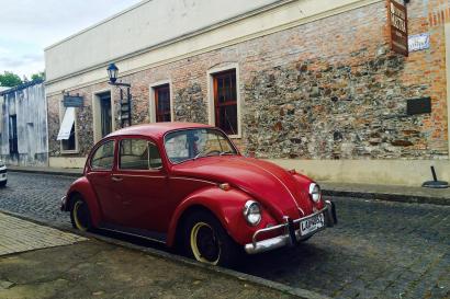 Red vintage Volkswagen Beetle parked on cobblestone street by a brick building. This alt text was added with Al; accuracy may vary.