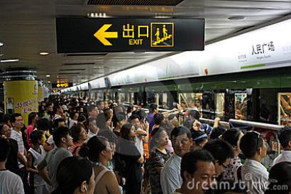 Crowded subway platform with people waiting for a train, exit sign visible above. This alt text was added with Al; accuracy may vary.