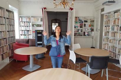 Girl standing in library with both hands palms up in a shrug, has a straight-faced expression