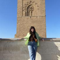A girl standing in front of the Tour Hassan, a brown tower with intricate detailing 
