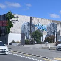 Image features a city street with cars on it, in the background is a large building with a mural on the side of it featuring two adult elephants and two young elephants.