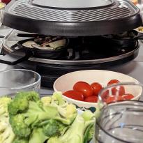 Table with bowl of tomatoes and broccoli in front of a raclette grill. 
