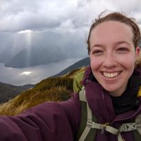 A selfie of me with my pack on and clouds shooting down rays of light in the background onto a lake. 