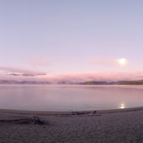 A panoramic view of a lake at dawn with the moon rising over it and mist rising off the water 