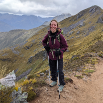 A photo of me with my pack and hiking poles standing on a ridge. The track is visible on a ridge in the background, along with sweeping mountains and white clouds. 