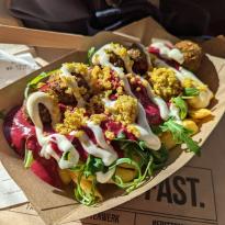 Cardboard plate with poutine with small balls of falafel scattered in it, covered in pink humus and yogurt dressign with arugula