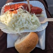 White cardboard plate with Currywurst and Krautsalat on it, bread roll on napkin on the side