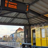 Subway station with yellow subway car with door open on the bottom right, top left corner shows sign with U3 Krumme Lanke and U1 Uhlandstrasse on it, in the background there's the Berlin skyline at twilight