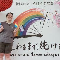 Author, Macks, standing in front of a poster from Tokyo Pride with mini pride flags.