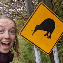 Me looking excited next to a yellow road sign with a silhouette of a Kiwi bird on it