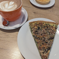 Yellow and green quiche with sesame seeds, an orange coffee cup filled with chai that has leaf latte art on top, and a round yellow custard lemon tart with blueberries on top all sit on a wooden table
