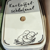White plastic box that has the words "Kartoffel schlafsaal" written on them, below the words are a drawing of two potatoes sleeping while cuddling each other 