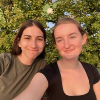 Two young women on a bench with trees behind it smiling at the camera
