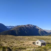 A small hut in a field of tussock on a mountain with the peaks of other mountains in the distance 