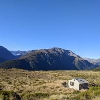 A small hut in a field of tussock on a mountain with the peaks of other mountains in the distance 