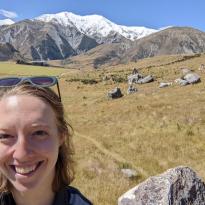 A selfie of me smiling in front of a big field with boulders and snowy mountains in the background